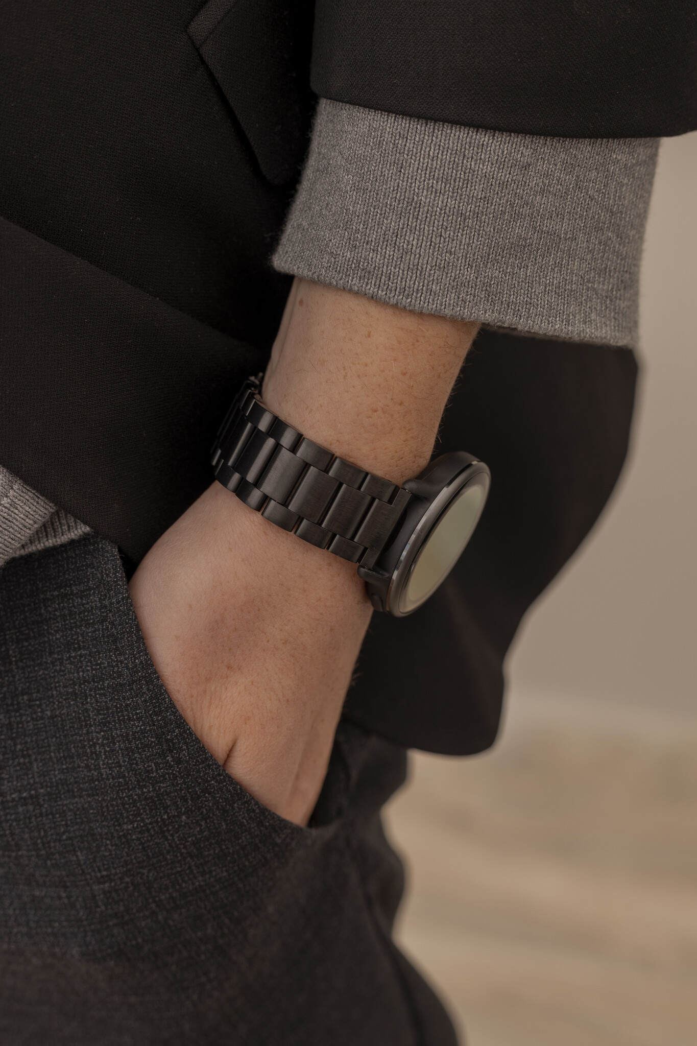 Das Handgelenk einer Person mit einer schwarzen Uhr und einem schwarzen Metallband. Die Person trägt ein graues Hemd und eine schwarze Jacke.