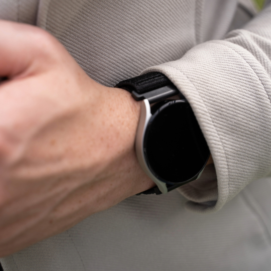 Das Handgelenk einer Person mit einer silbernen Smartwatch. Das Armband ist schwarz, sie trägt eine hellgraue Jacke.