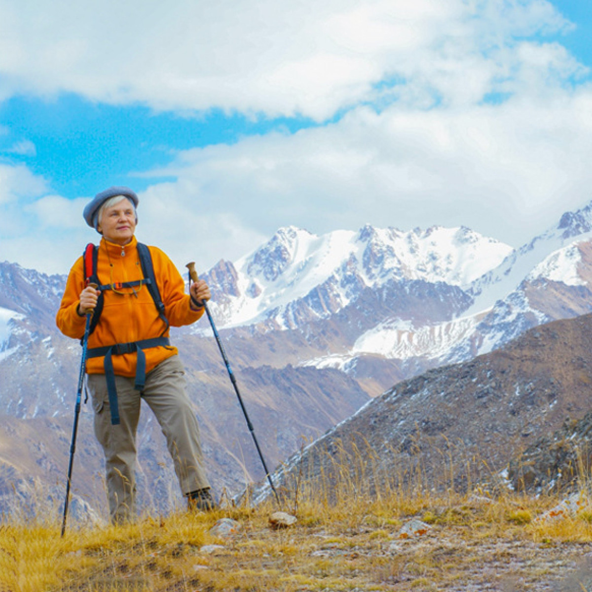 Starsza kobieta z kijkami do wędrówek na górze, w tle ośnieżone szczyty.