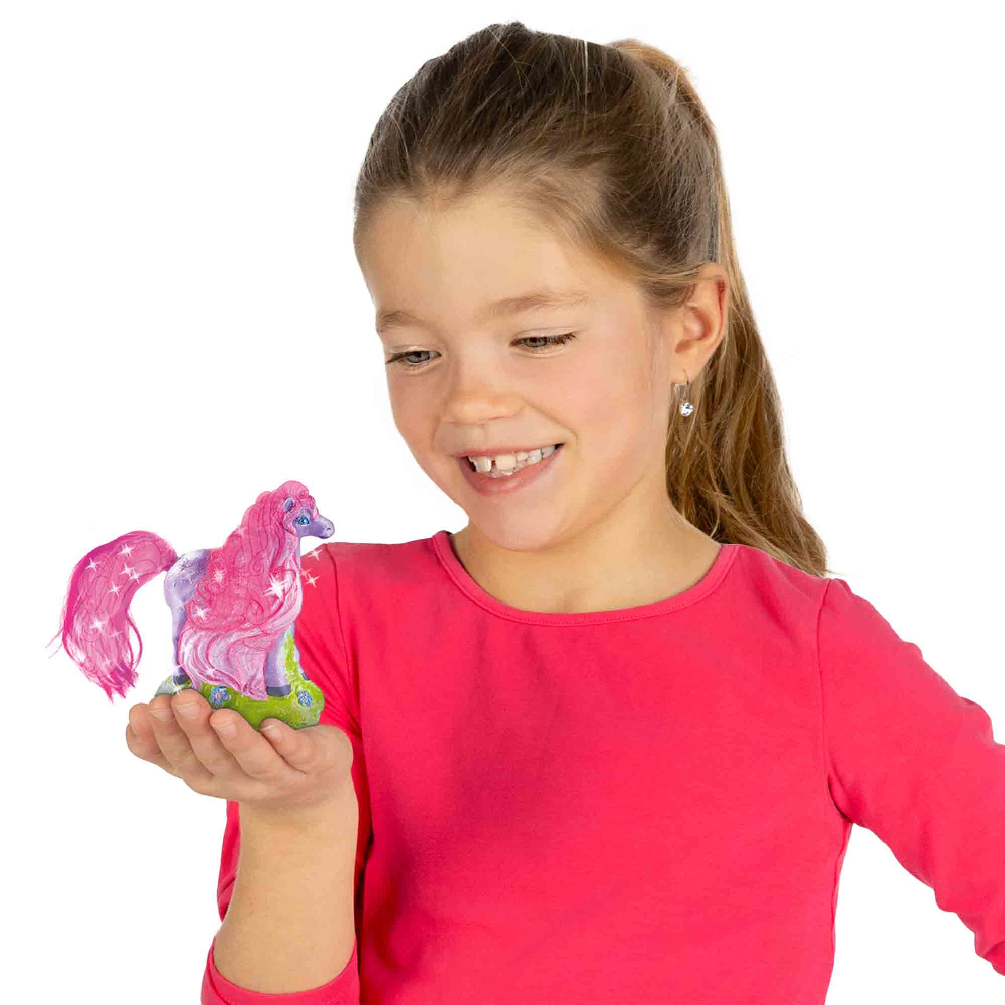 Dziewczyna trzymająca różową figurkę konia z błyszczącym ogonem na zielonej podstawie, uśmiechając się do kamery.