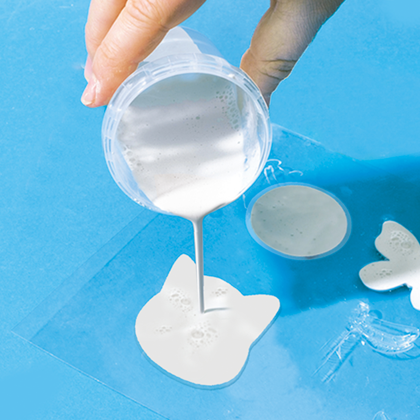 Ręka wylewa płyn do formy w kształcie kota na niebieskiej powierzchni.