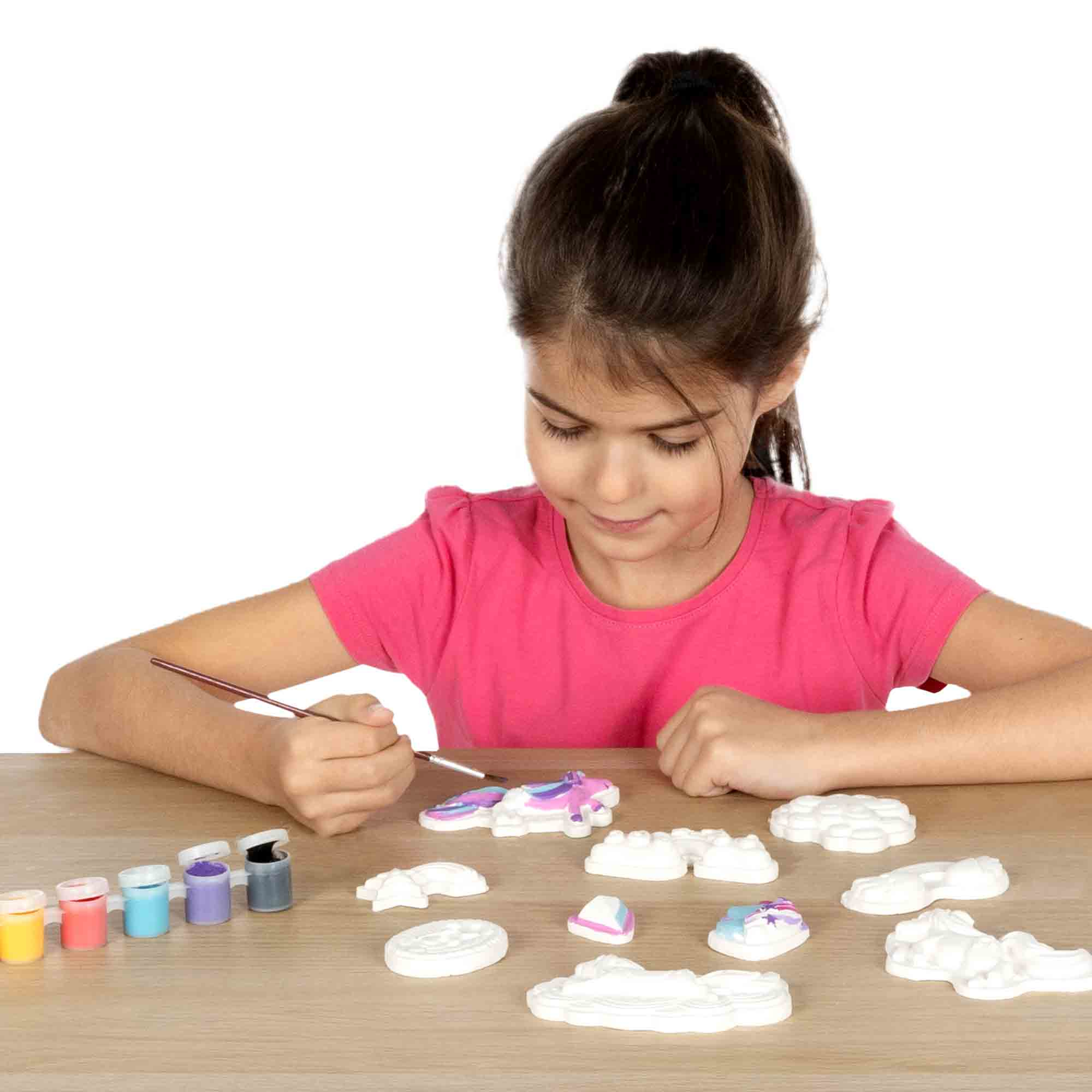 Dziewczynka maluje figurkę jednorożca na drewnianym stole. W pobliżu znajdują się różne kolory farb i figurki.