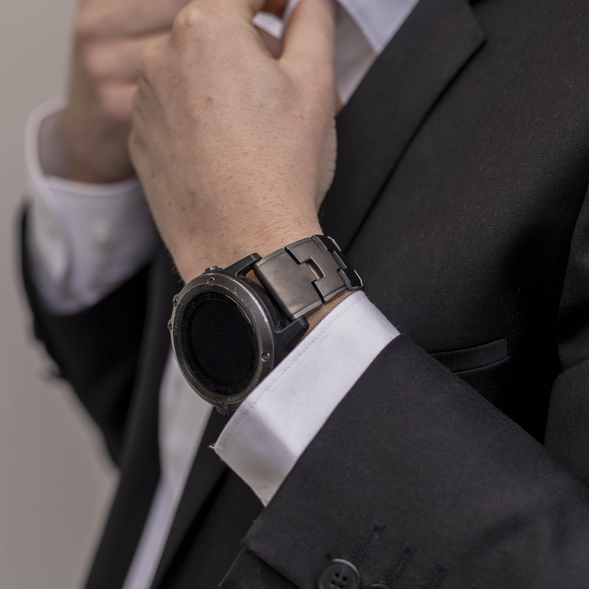 Person passt Uhr an, trägt Anzug, weißes Hemd. Schwarze Uhr am Handgelenk. Nahaufnahme, neutraler Hintergrund.
