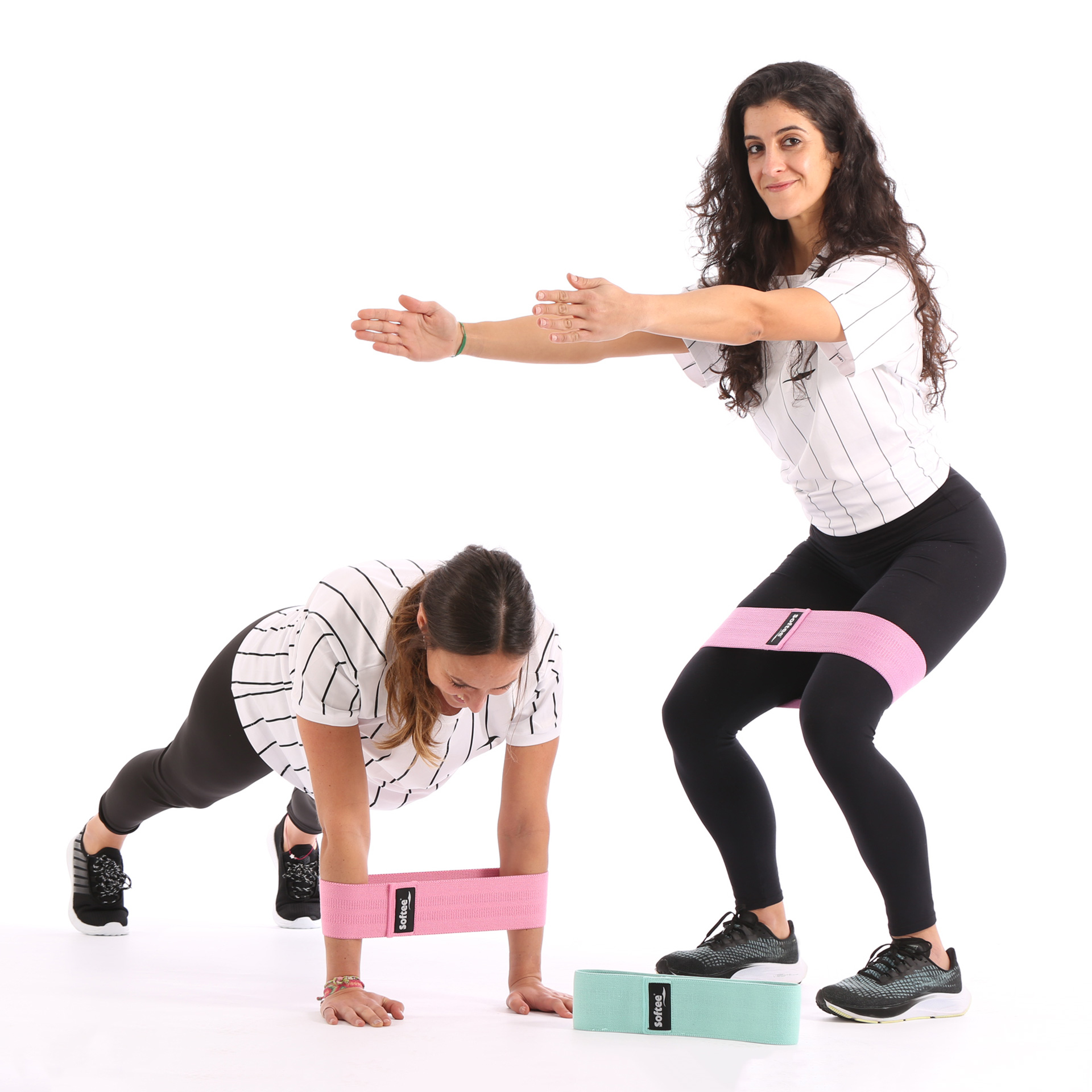 Dos mujeres haciendo ejercicio con bandas de resistencia, una haciendo sentadillas, la otra en plancha. Se ven bandas rosas y verdes.