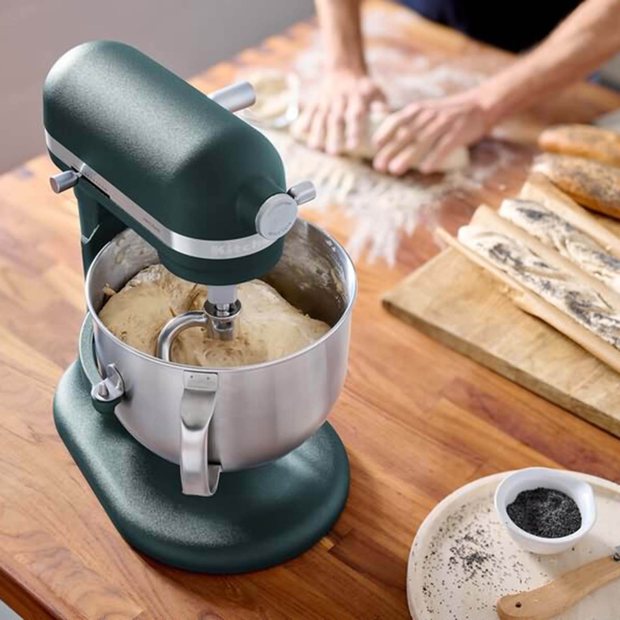 Grüner Küchenmixer mit Teig in der Schüssel, Holztisch und Person, die im Hintergrund knetet.