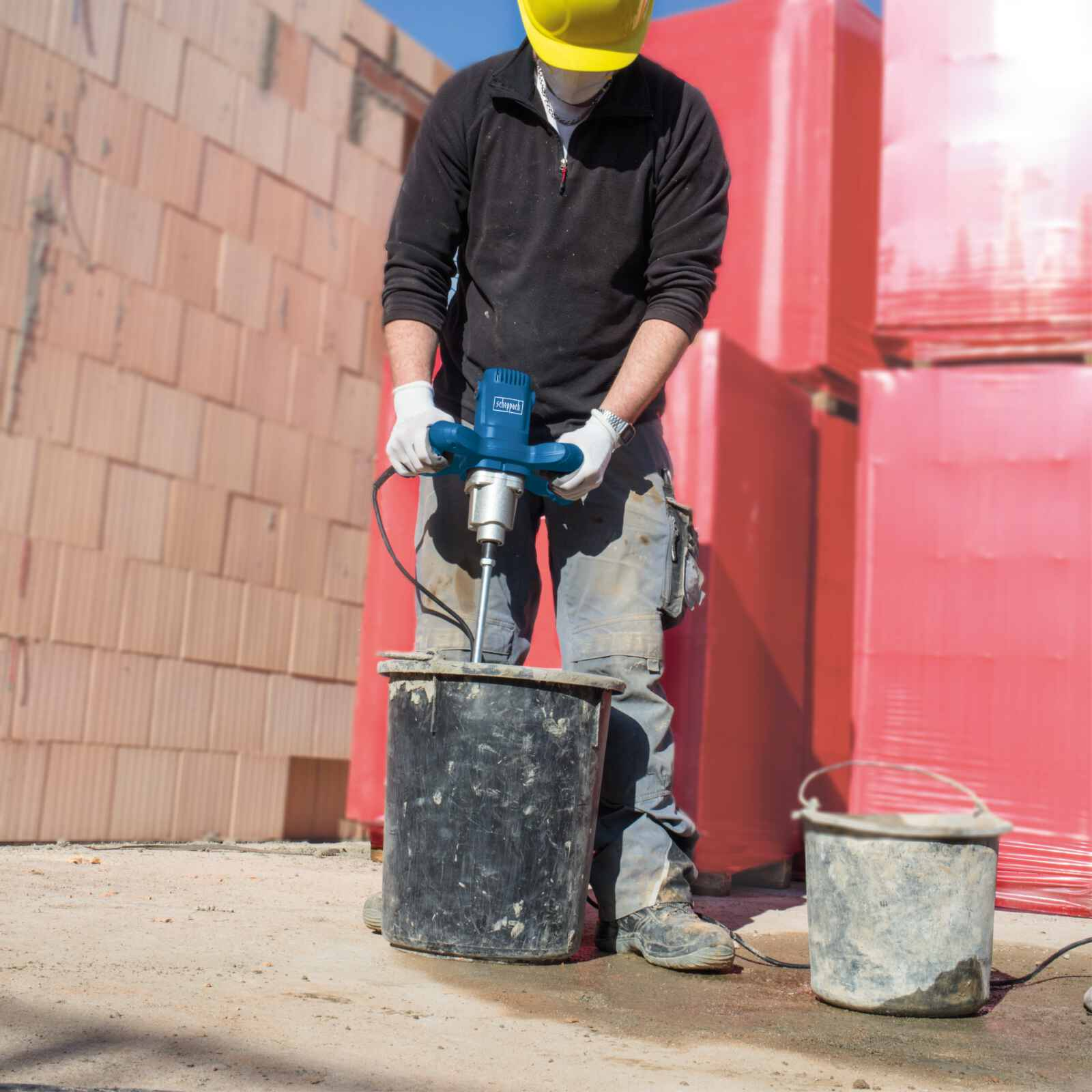 Uomo con casco giallo usa miscelatore per mescolare cemento in un secchio nero, sfondo cantiere.