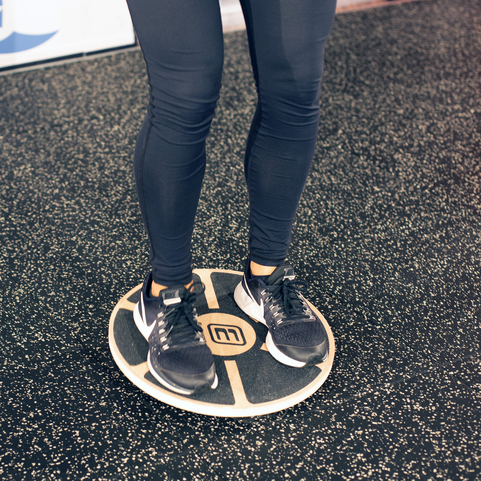 Persona con leggings y zapatos negros en tabla de equilibrio, blanco y negro, en suelo gimnasio.