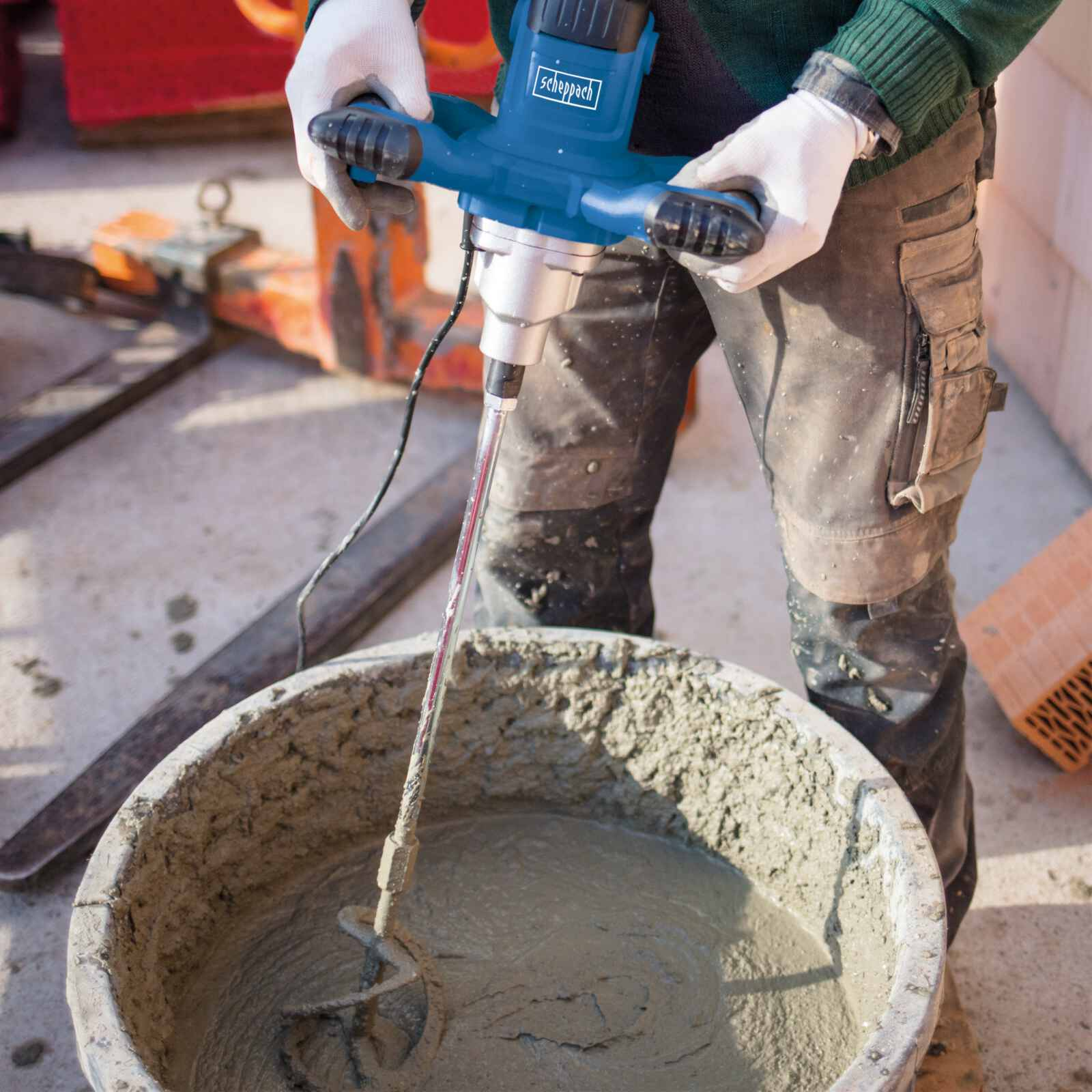Persona che mescola cemento in un secchio con un miscelatore blu. Indossa guanti.
