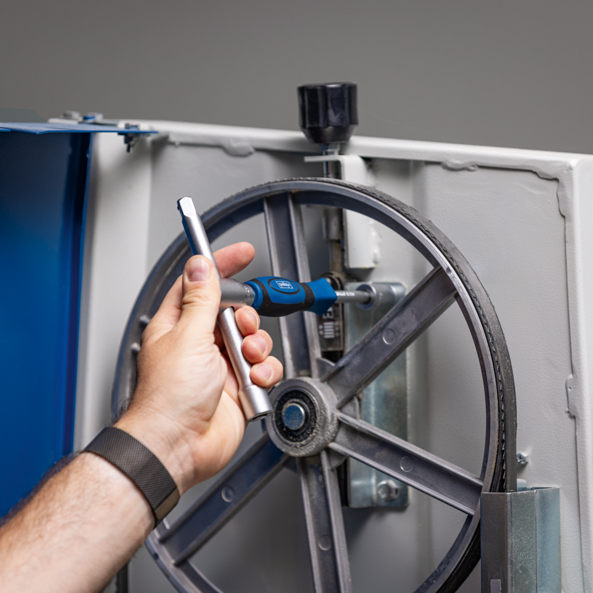 A person's hand using a tool to adjust a machine's wheel. The tool is blue and silver.