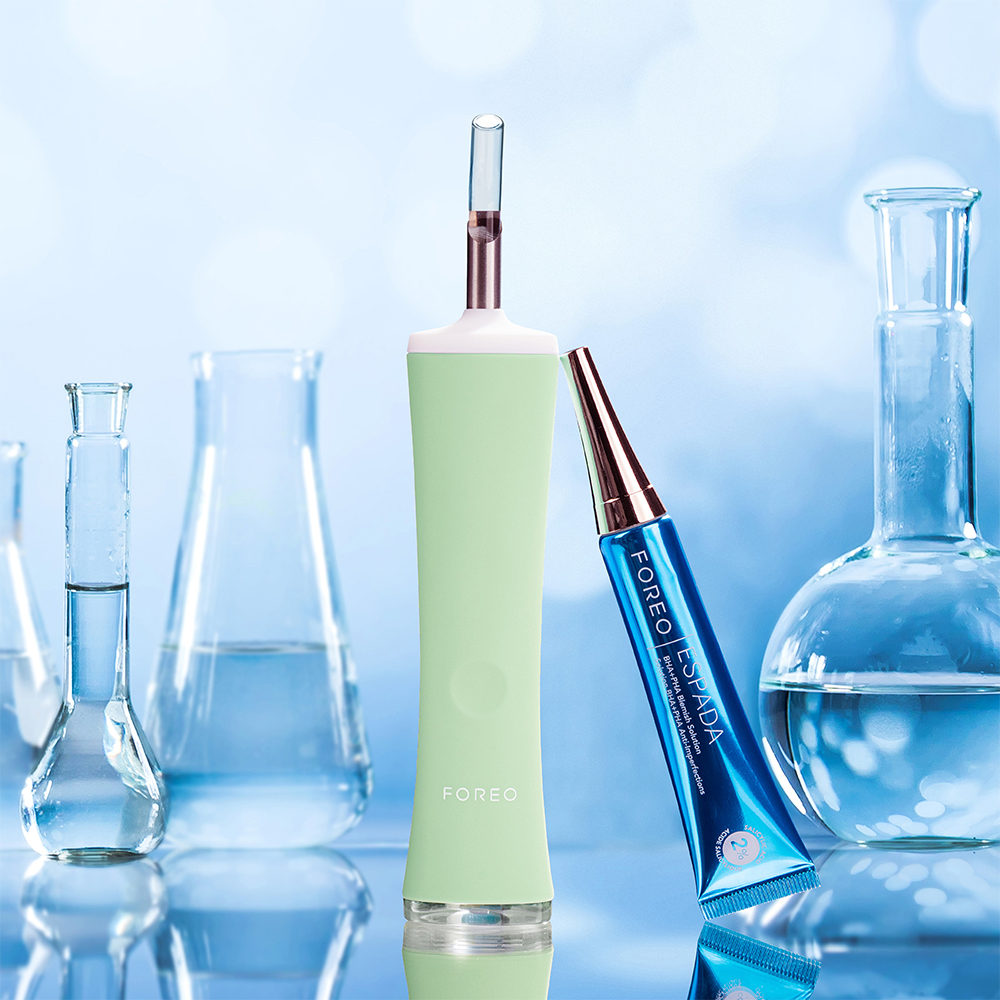 Green Foreo device and blue tube with a pink tip are on a glossy surface with lab equipment in the background.