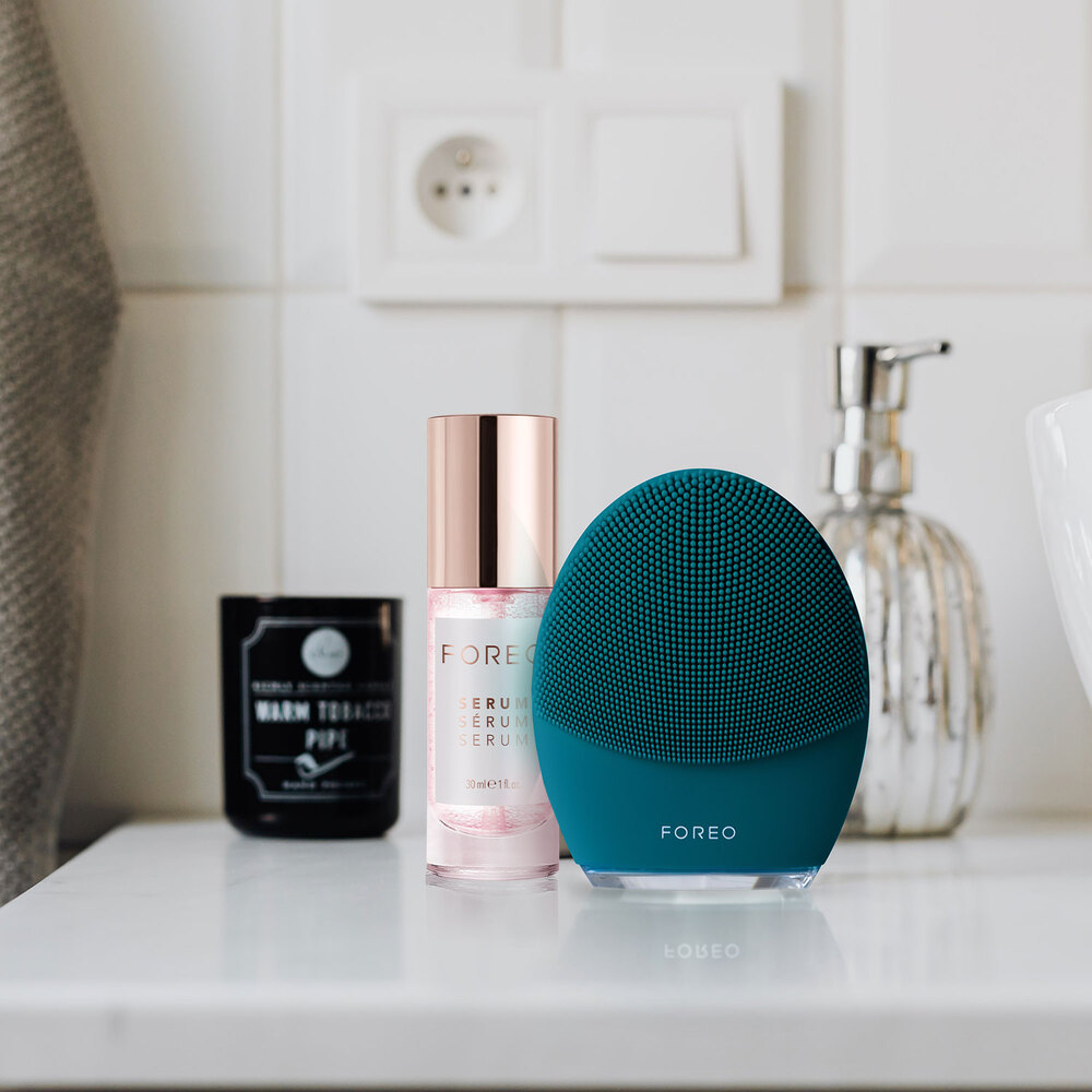 Een Foreo gezichtsreinigingsapparaat, een serumfles en een kaars op een wit aanrecht in een badkamer.