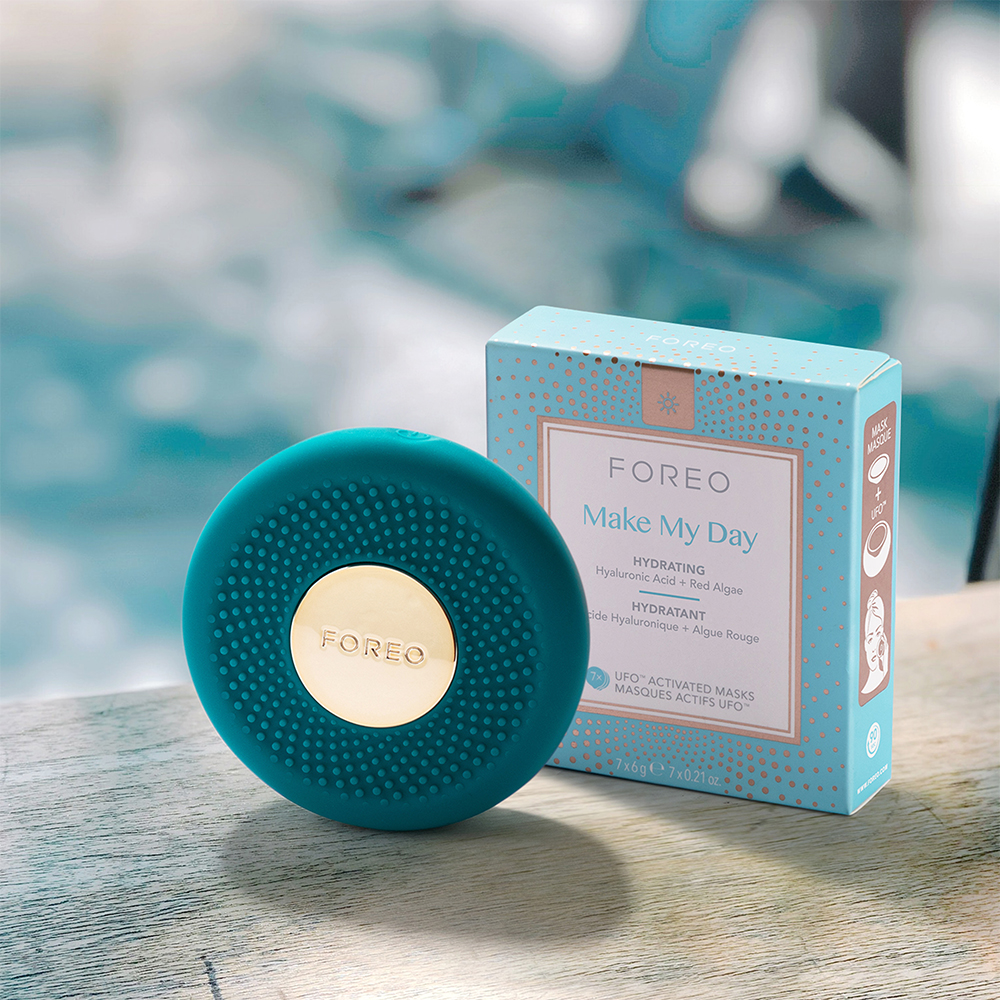 Een turquoise huidverzorgingsapparaat naast een huidverzorgingsproductdoos op een houten oppervlak bij water.