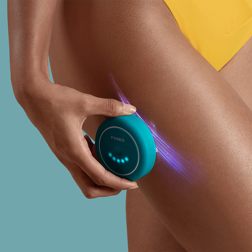 A woman is using a turquoise Foreo device on her thigh.