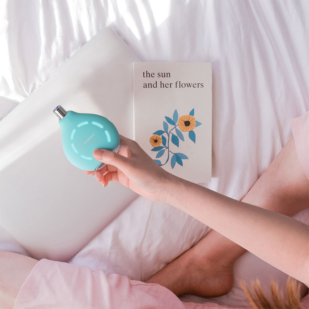 Femme tenant un appareil Foreo turquoise à côté d'un livre sur un lit.