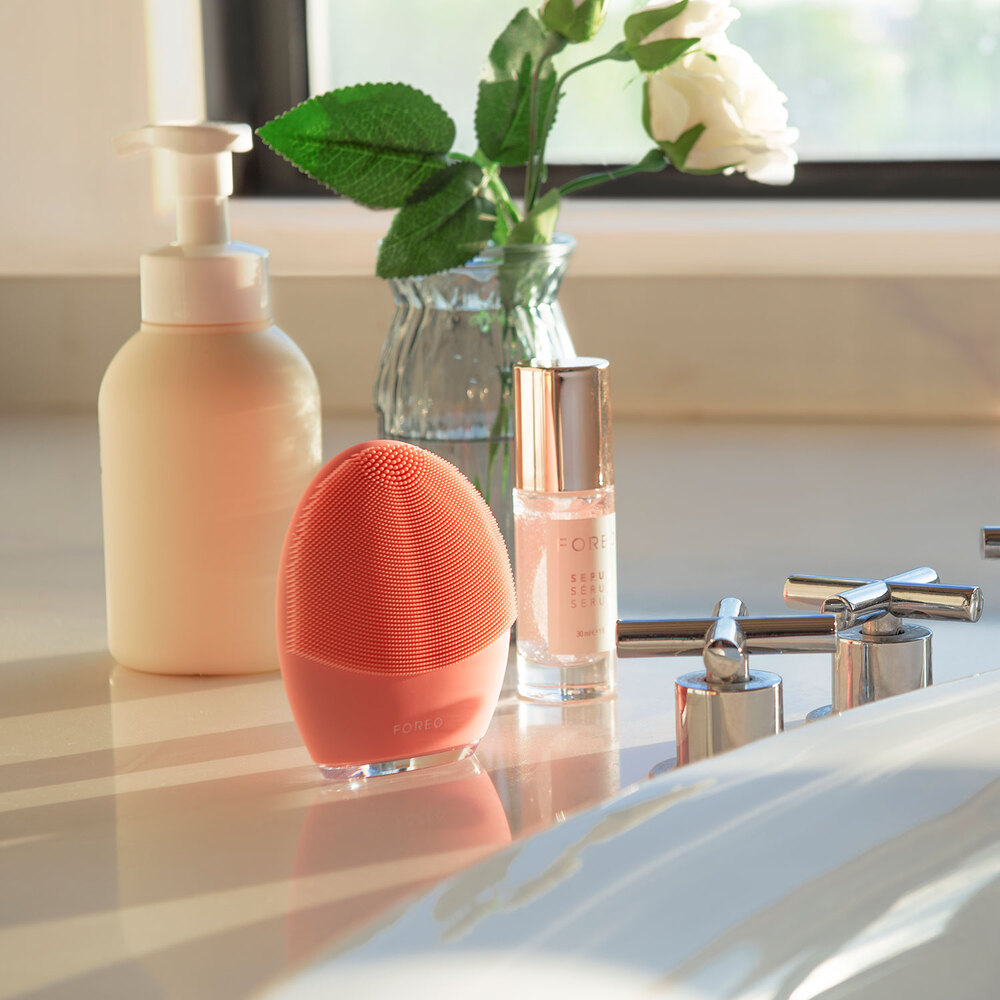 A Foreo Luna, serum, and lotion on a counter. A vase with flowers is behind them.