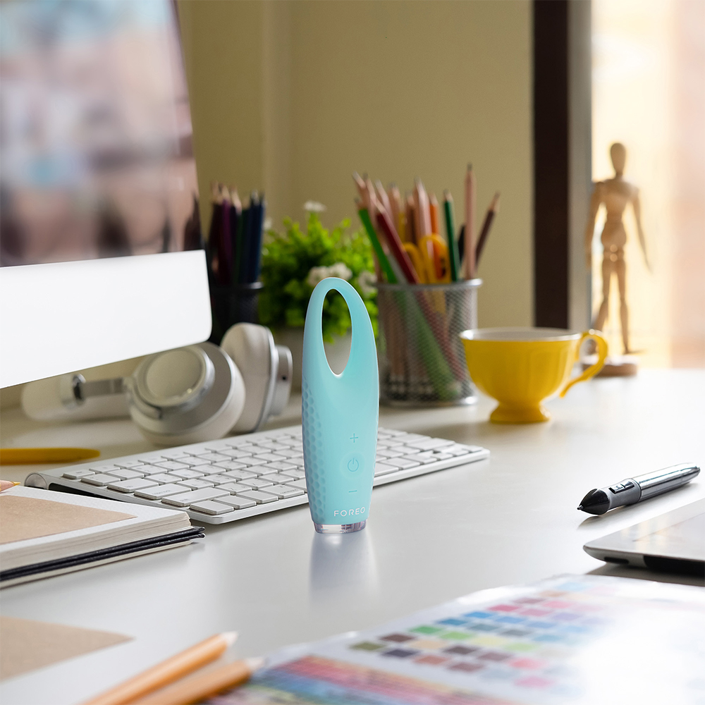 A blue Foreo Iris 2 eye massager on a desk. A keyboard, headphones, and other items are also present.