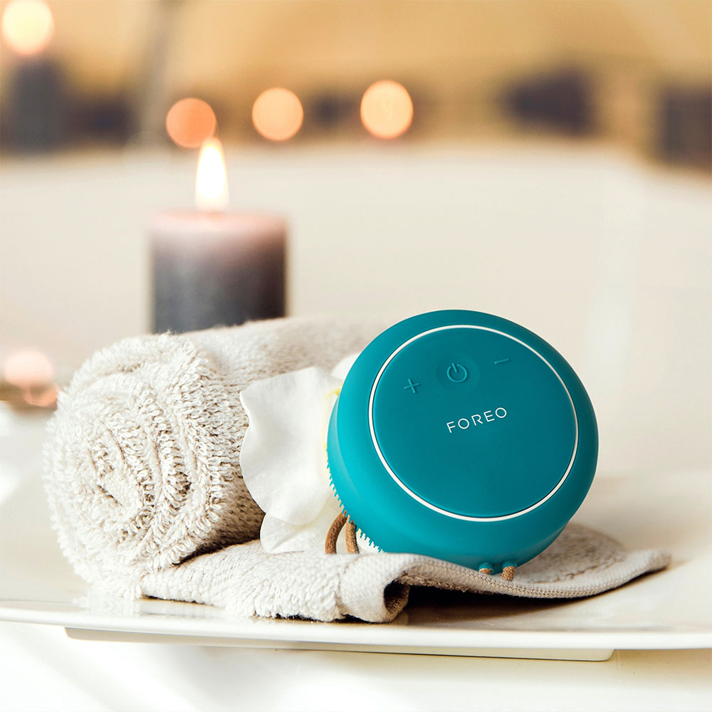 Een turquoise Foreo-apparaat, een handdoek en een bloem op een wit bord bij een brandende kaars.