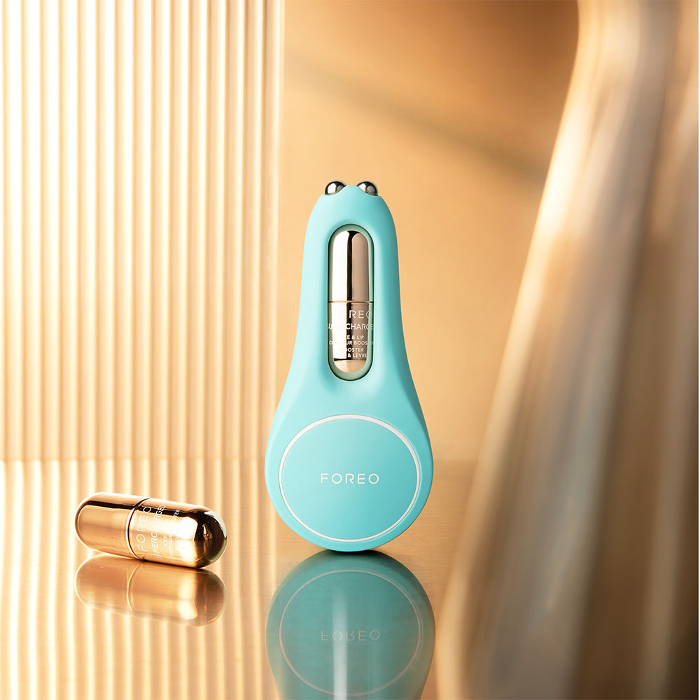 A teal Foreo device and a gold capsule, on a reflective surface, with a golden backdrop.