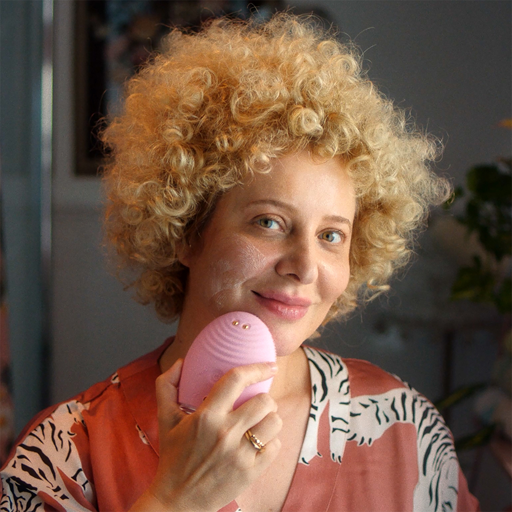Woman with curly hair using a pink facial device. She is smiling. Background is blurred.