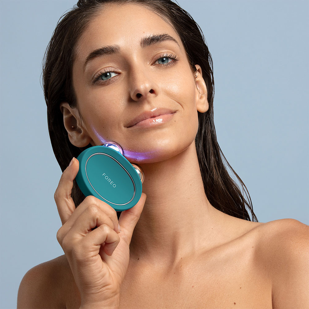 Woman uses teal FOREO device on her face. Blue background. Product shown is FOREO.