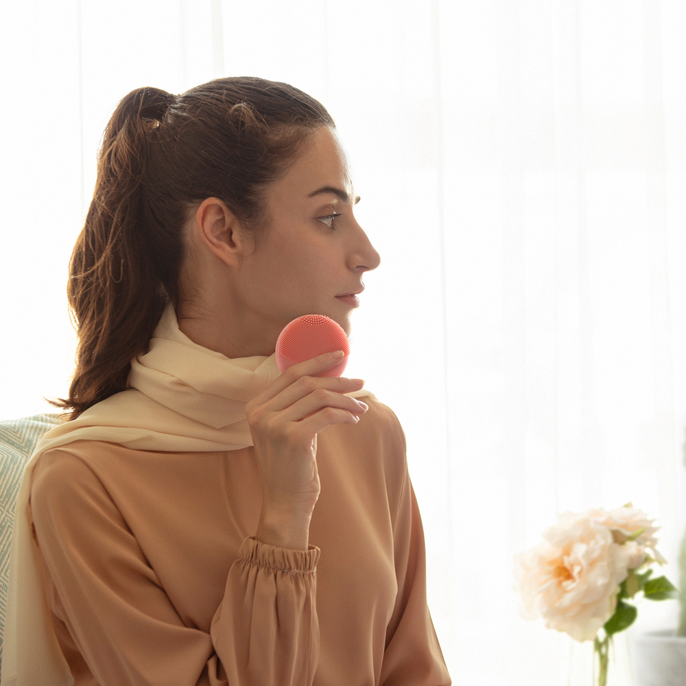 A woman with a beige scarf and top holds a pink FOREO device to her face. White background.