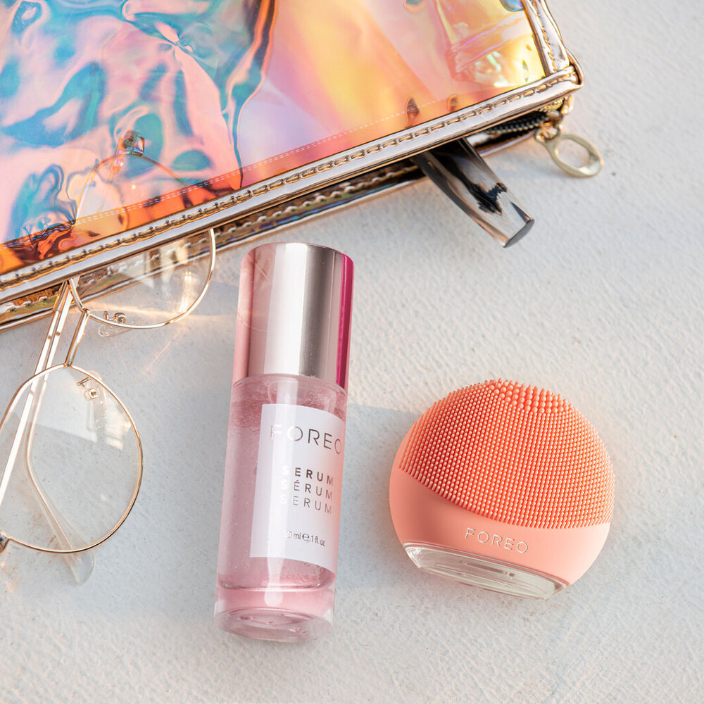 A Foreo serum bottle, a face brush, a makeup bag, and glasses on a white surface.