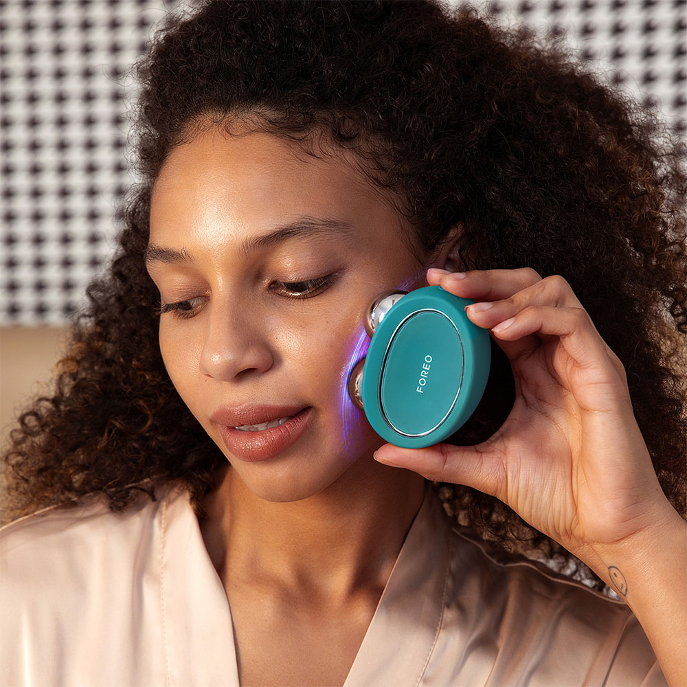 Une personne tenant un appareil Foreo turquoise près de son visage, avec un fond beige.