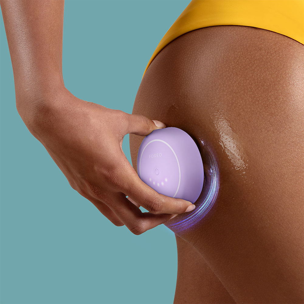 Une main utilise un appareil Foreo sur sa cuisse, avec un fond bleu.