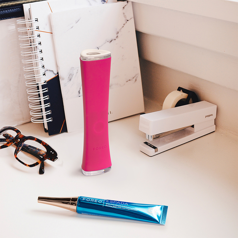A pink Foreo Espada 2 device, stapler, glasses, and other items on a white desk.