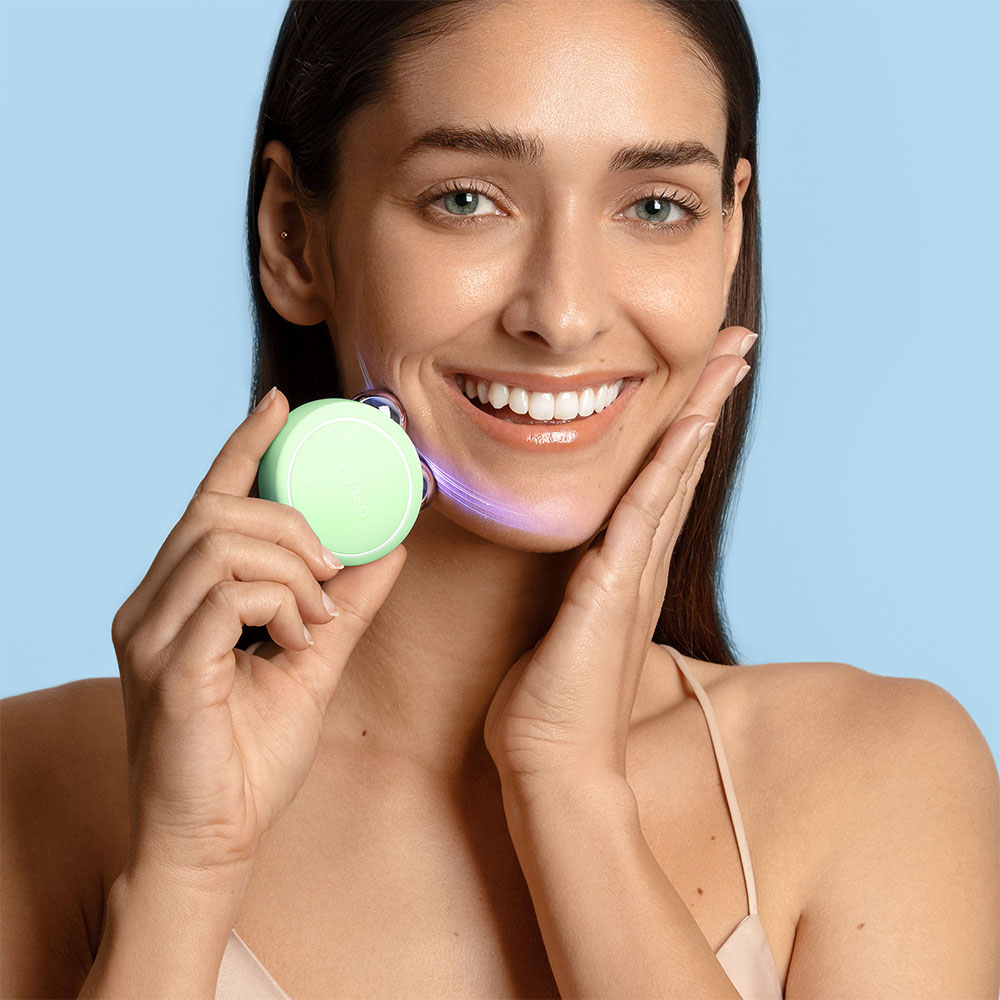 Woman holding a green Foreo device. She smiles and touches her face.