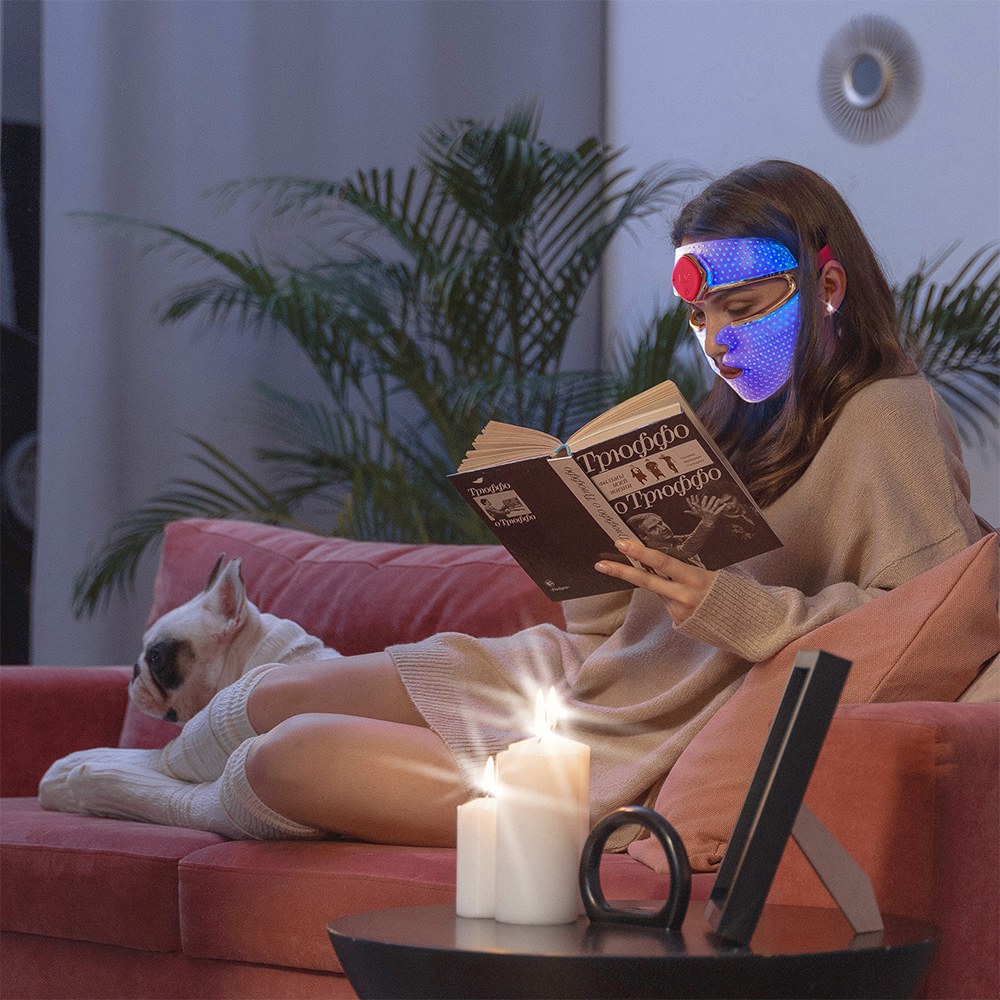 Femme lisant un livre sur un canapé avec un chien, un masque facial à lumière bleue sur son visage.