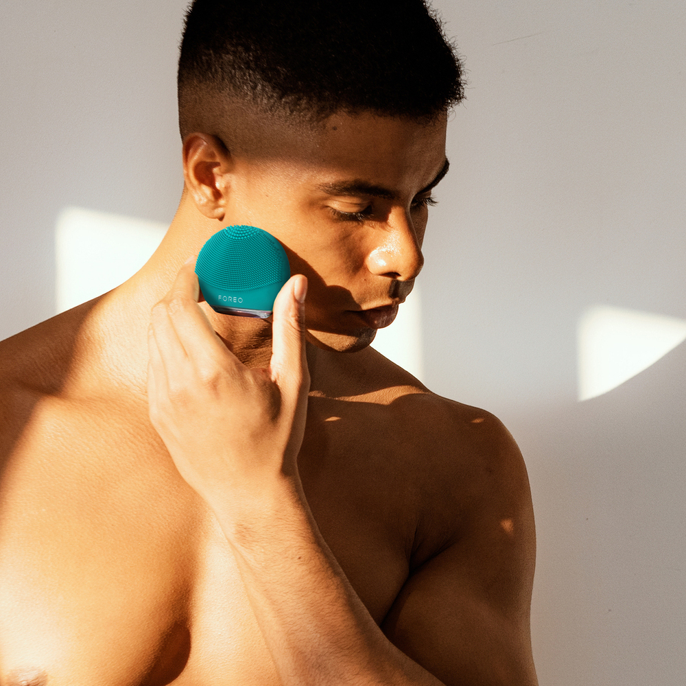Man using teal facial device on his cheek. White background.