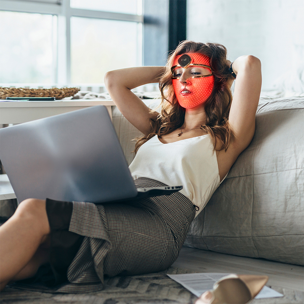 Woman wearing a red LED face mask, sitting near a laptop on a floor.