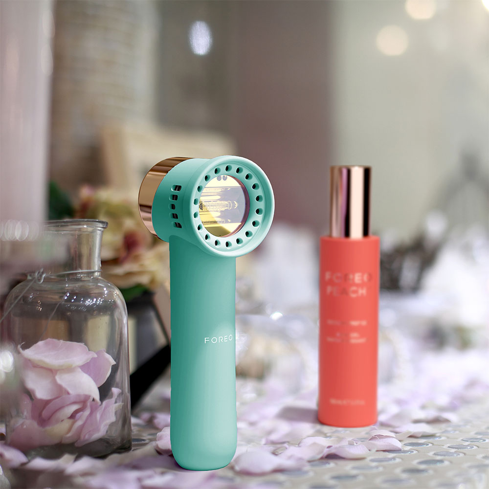 A teal and gold device and a coral-colored bottle on a surface, with rose petals.