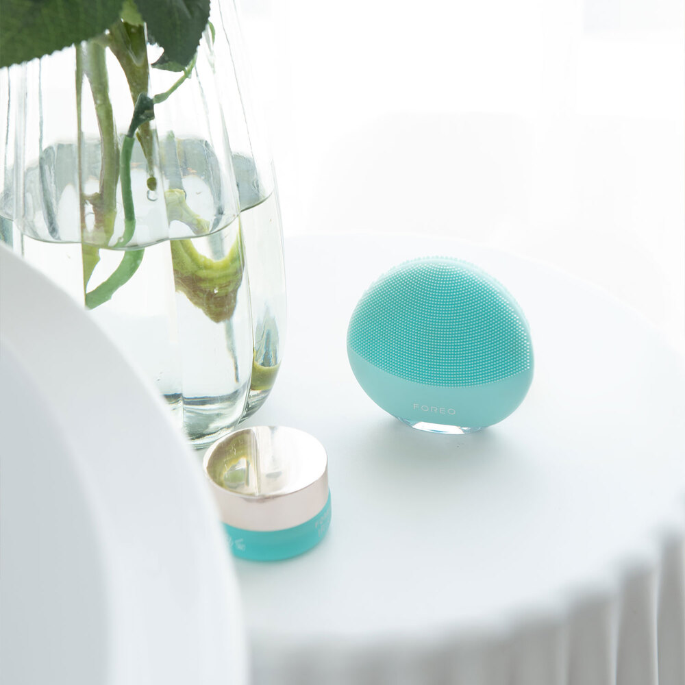 A teal facial cleansing device and skincare products on a white table, near a vase with water and flowers.