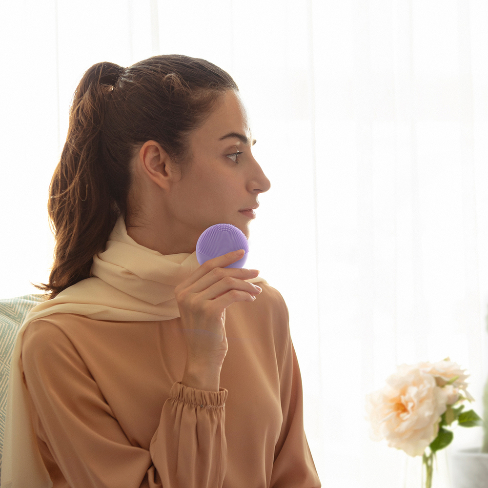 A woman holds a purple Foreo device near her face. She is looking to the side.