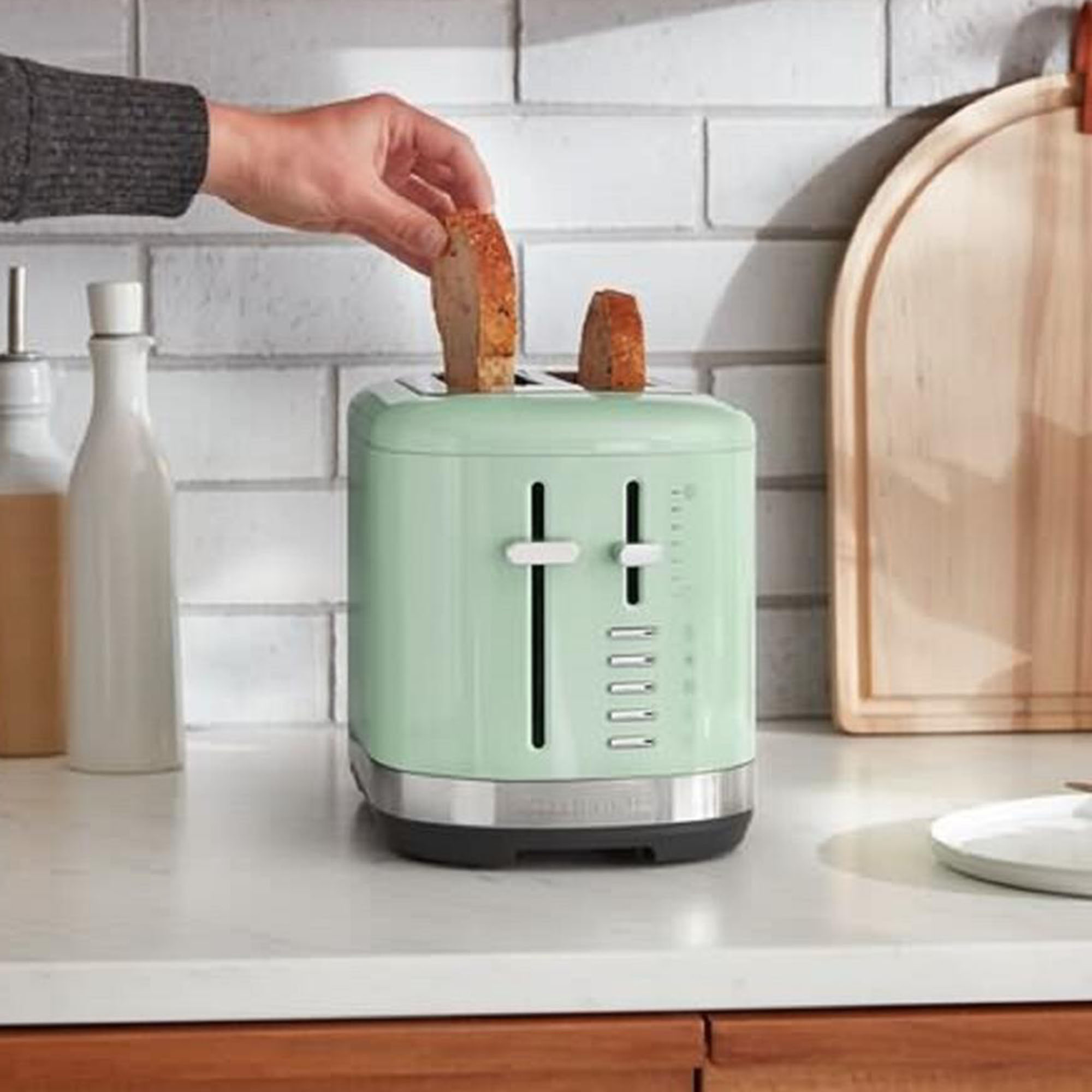 A person placing toast into a green toaster on a kitchen counter, with other kitchen items in the background.