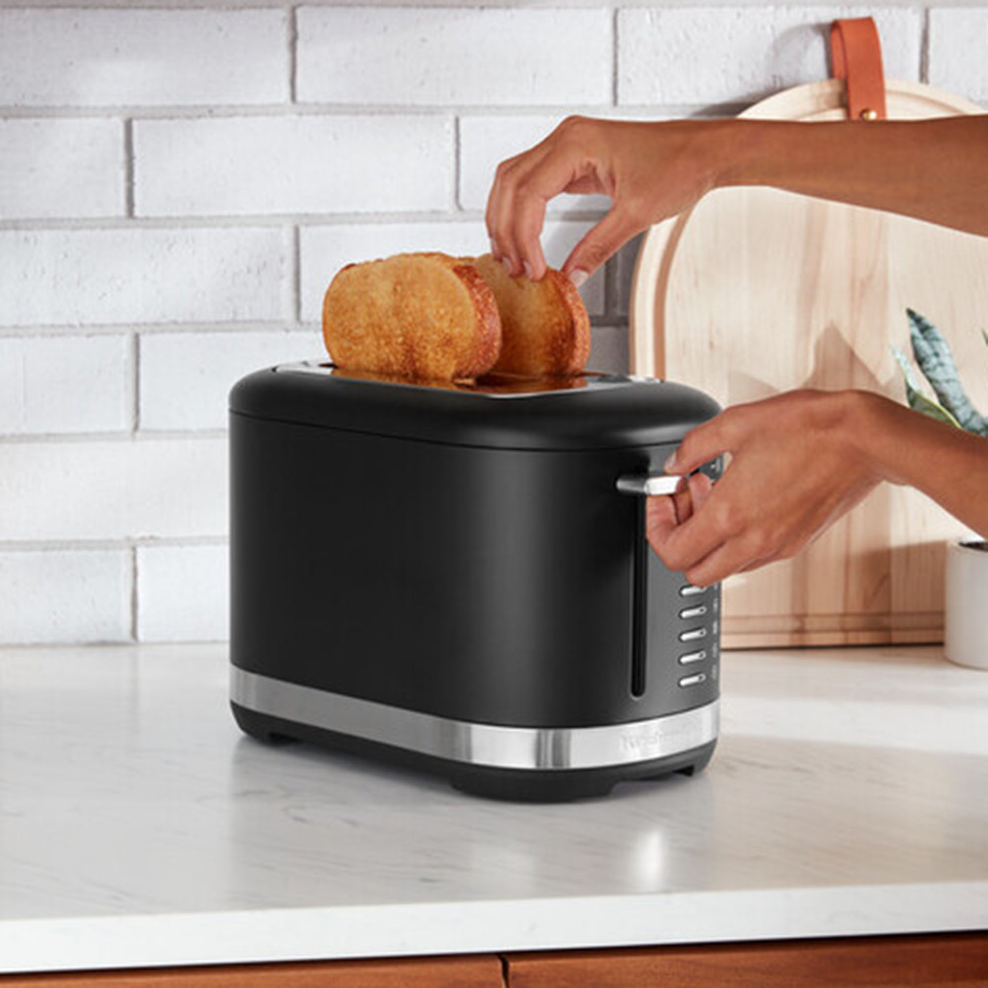 A person's hands placing toast into a black toaster on a kitchen counter, with other items.