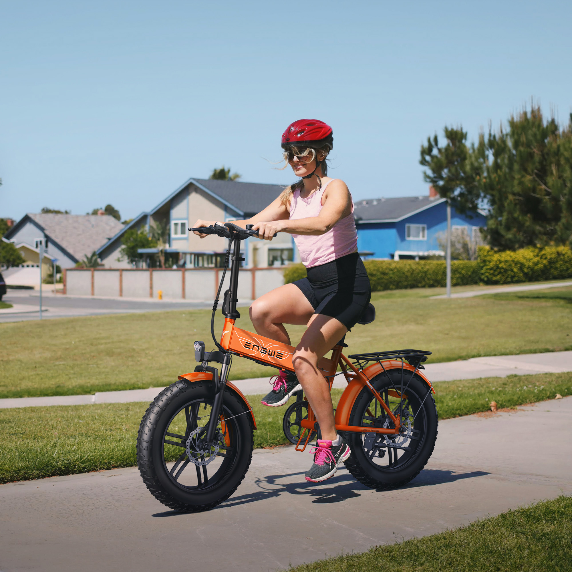 Mujer en top rosa y shorts negros montando bicicleta eléctrica naranja en camino pavimentado, día soleado.