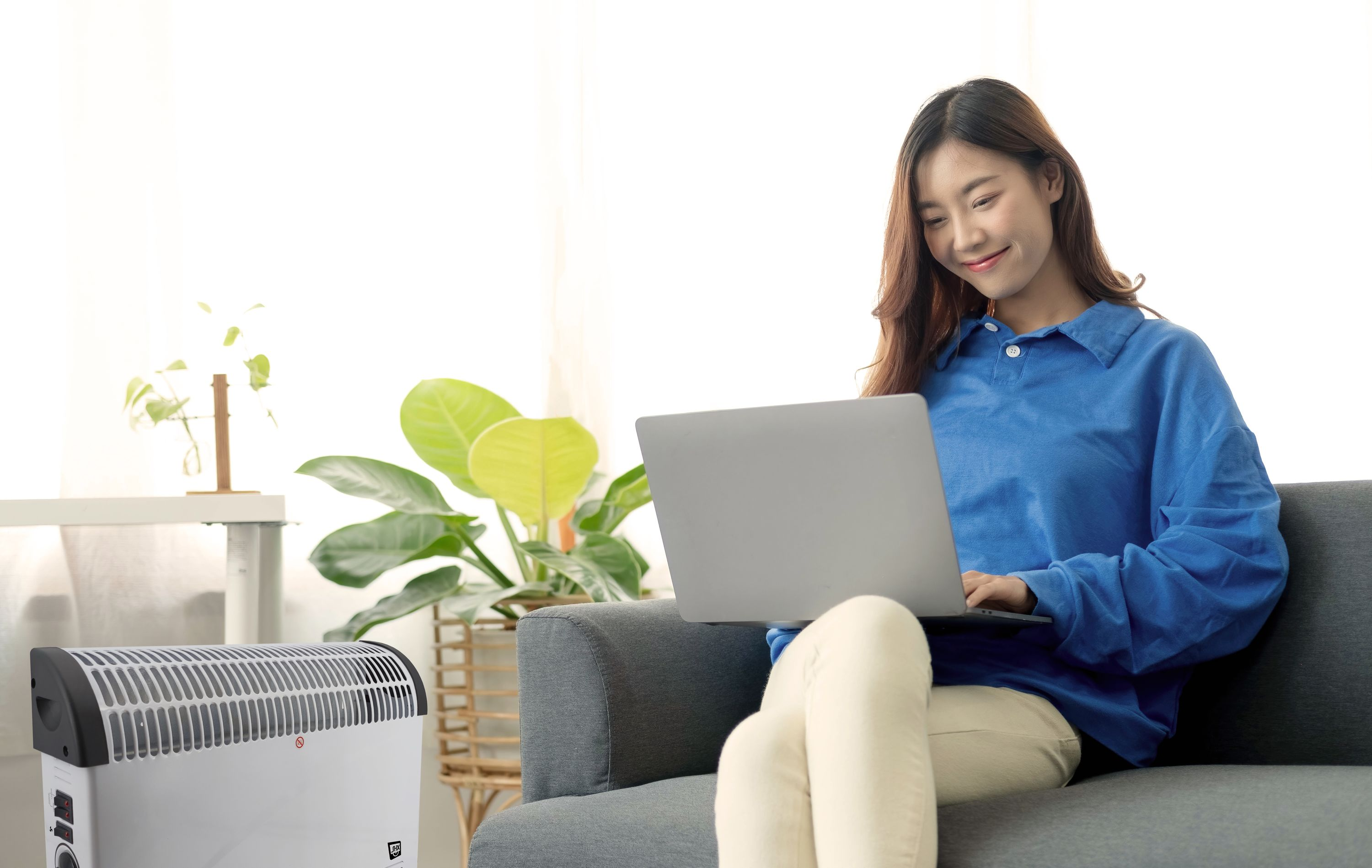 Kobieta korzystająca z laptopa na kanapie. Po lewej stronie znajduje się grzejnik. Roślina doniczkowa w tle.