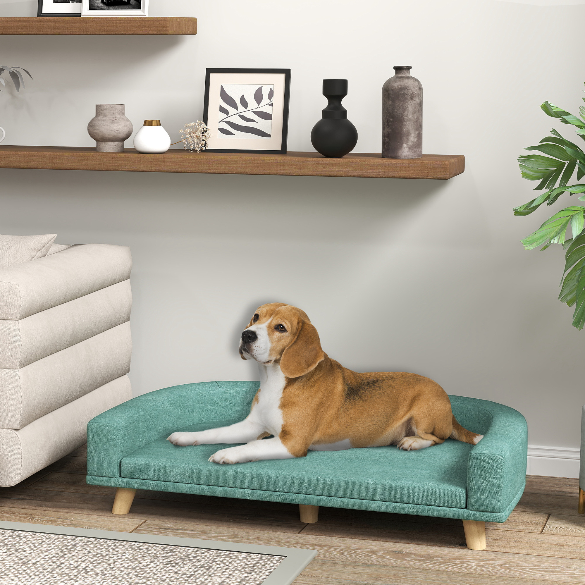 Un beagle yace en una cama para perros color verde azulado. Cerca, una mesa auxiliar con un jarrón y libros.