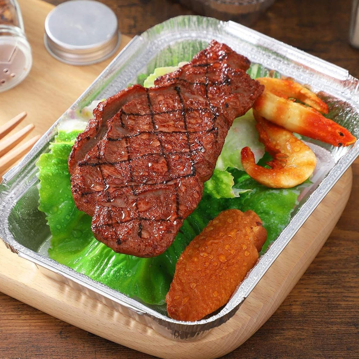 Gegrilltes Steak, Garnelen und eine Flügel in einem Folienbehälter auf Holz. Grüner Salat.