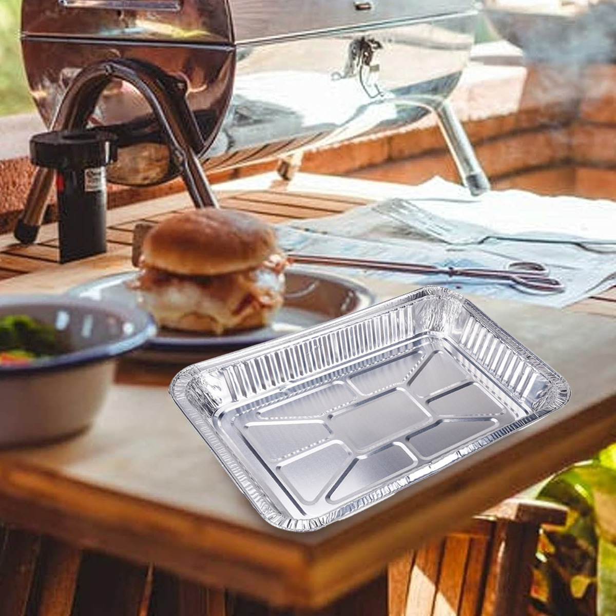 Alufolien-Tablett auf einem Holztisch, in der Nähe eines Grills und Burgers. Weißer Hintergrund.