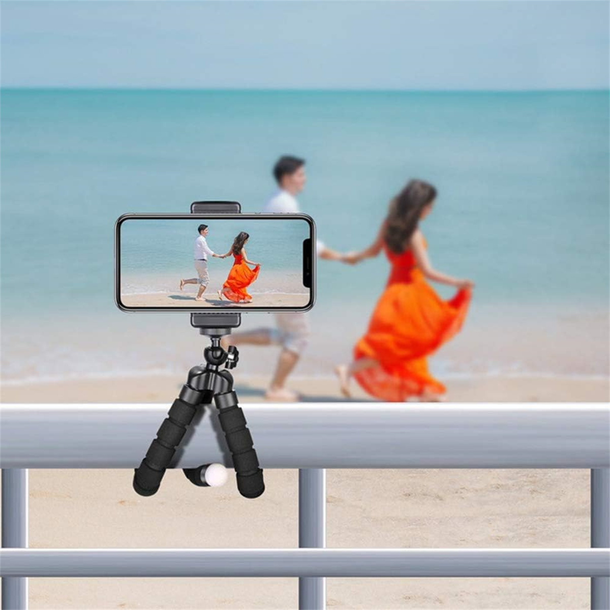 Ein Telefon auf einem Stativ zeigt ein Paar, das am Strand läuft. Der Hintergrund ist verschwommenes Meer und Himmel.