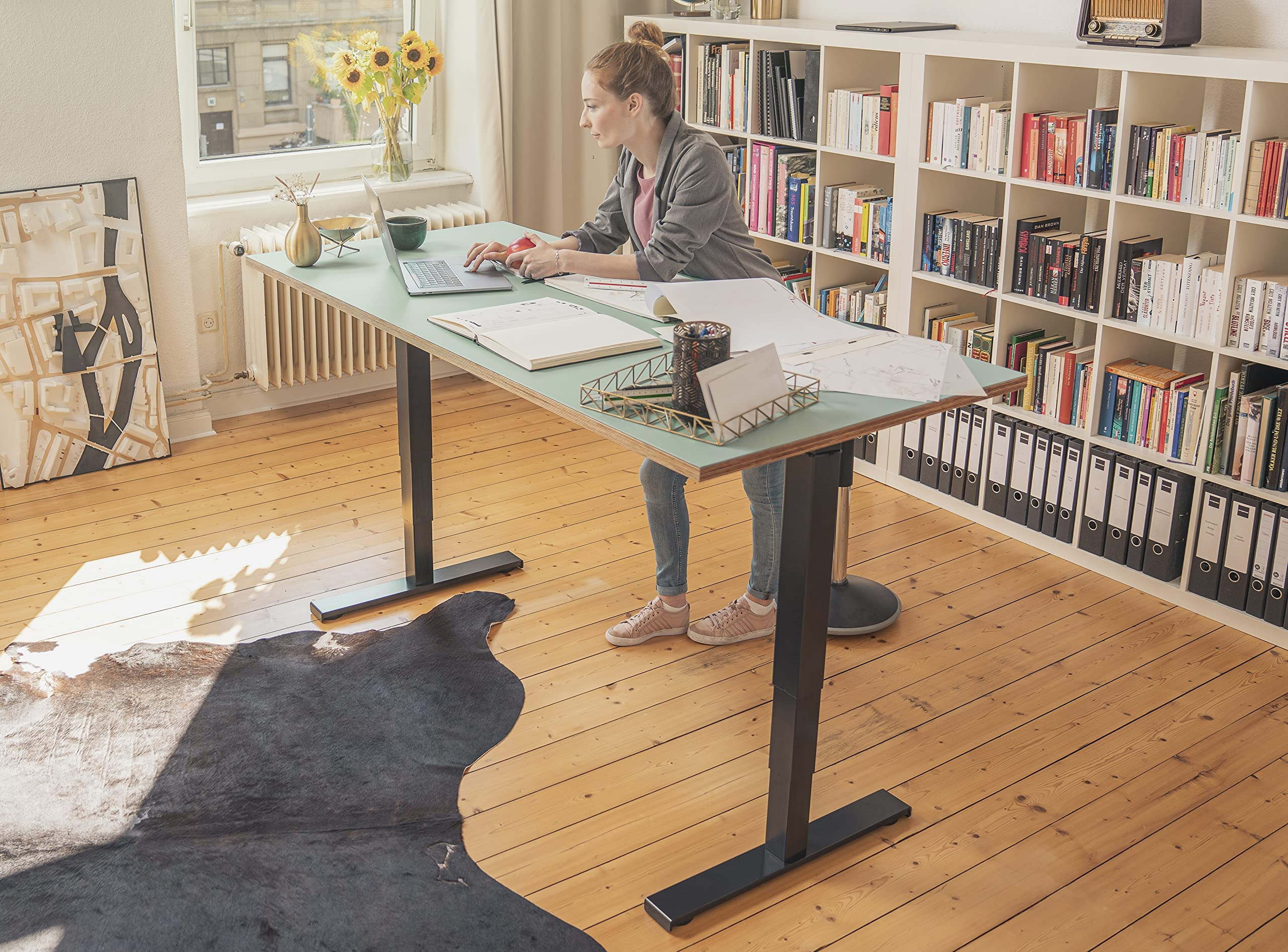Frau arbeitet an einem Schreibtisch mit Laptop. Bücherregale und ein Teppich im Hintergrund.