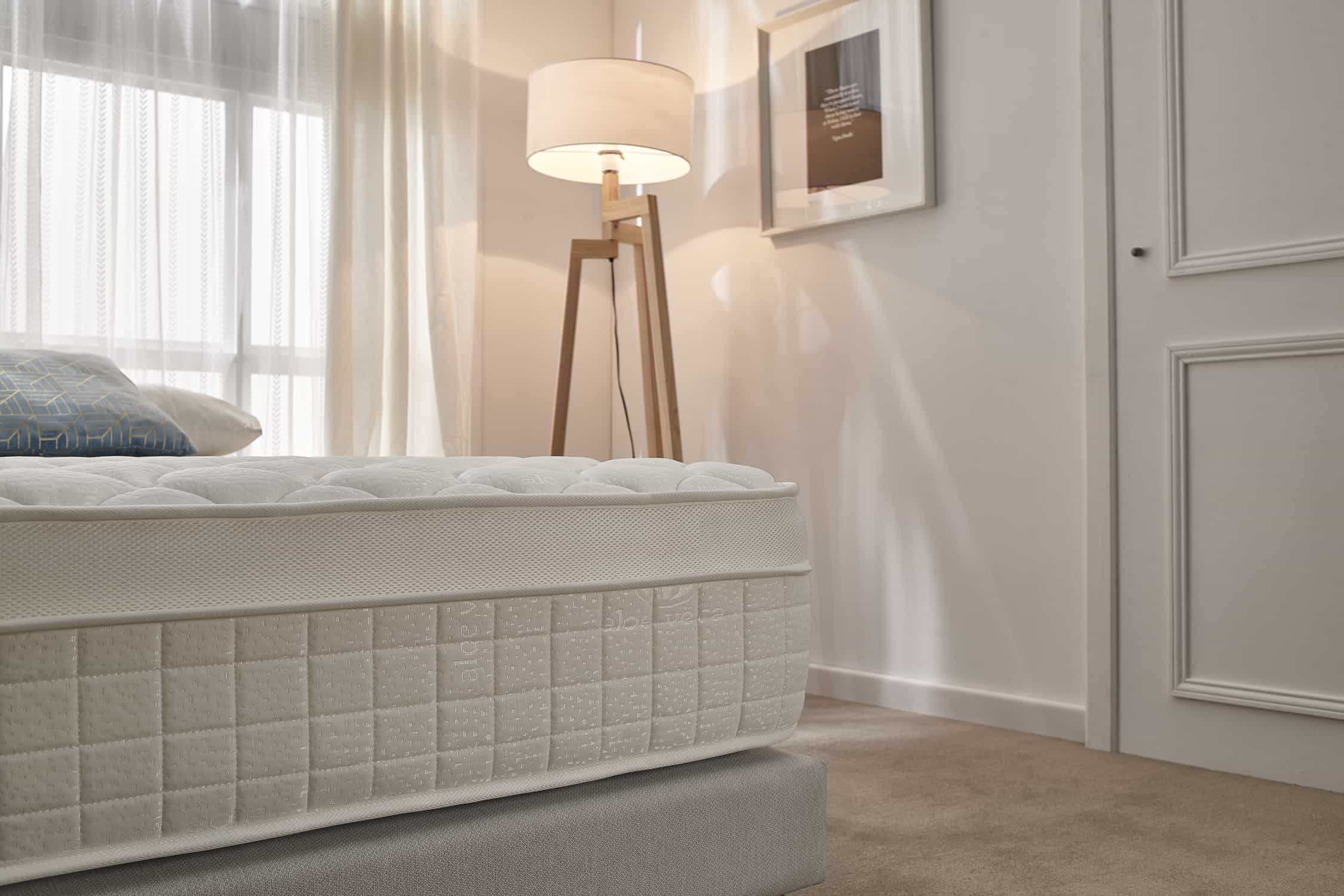 Dormitorio con cama. La cama tiene colchón blanco y base gris claro. Se ven una lámpara y una puerta.