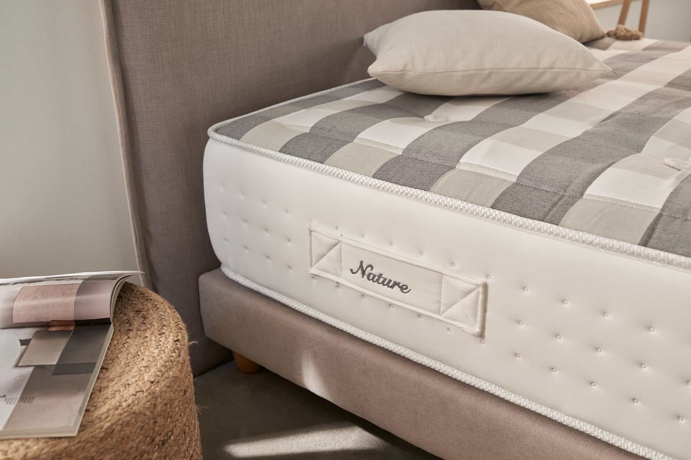 Primer plano de una cama con un colchón etiquetado como "Nature". Patrón a cuadros gris y blanco. Cabecero beige.
