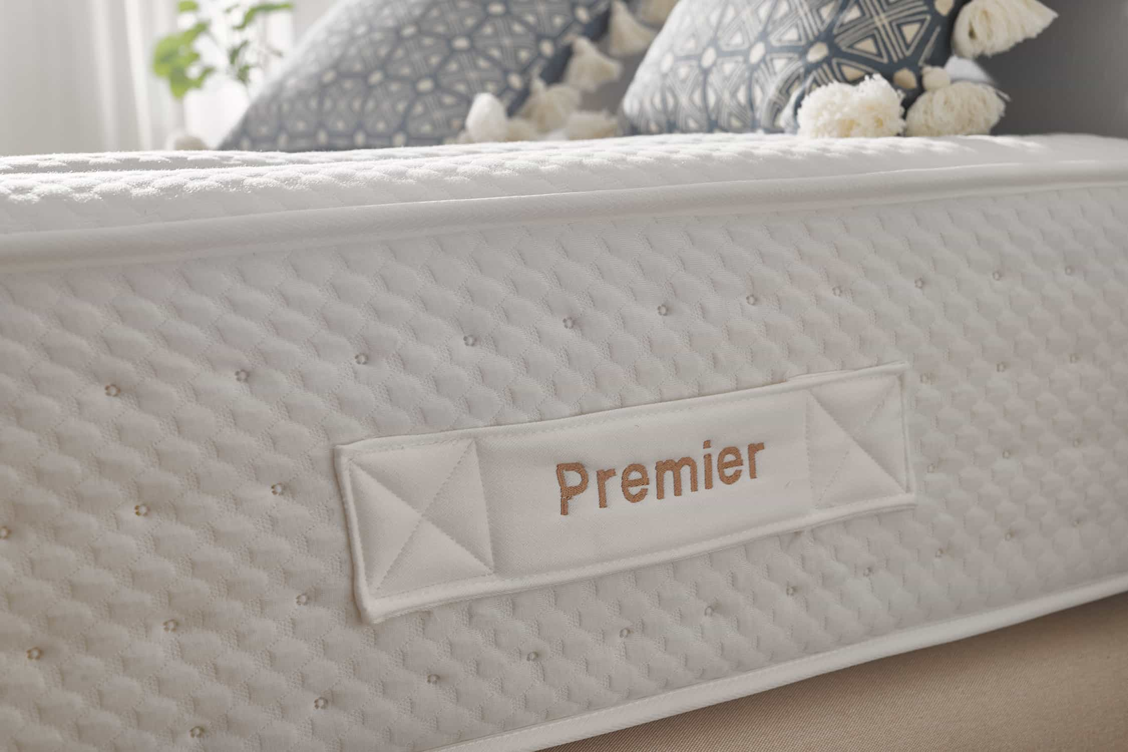 Colchón blanco con la palabra 'Premier', colocado en una cama con almohadas y objetos decorativos al fondo.