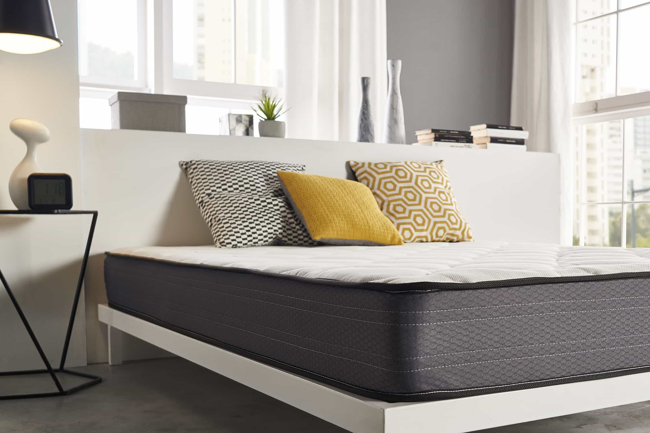 Una cama con colchón, almohadas y una lámpara, contra una pared blanca con elementos decorativos.