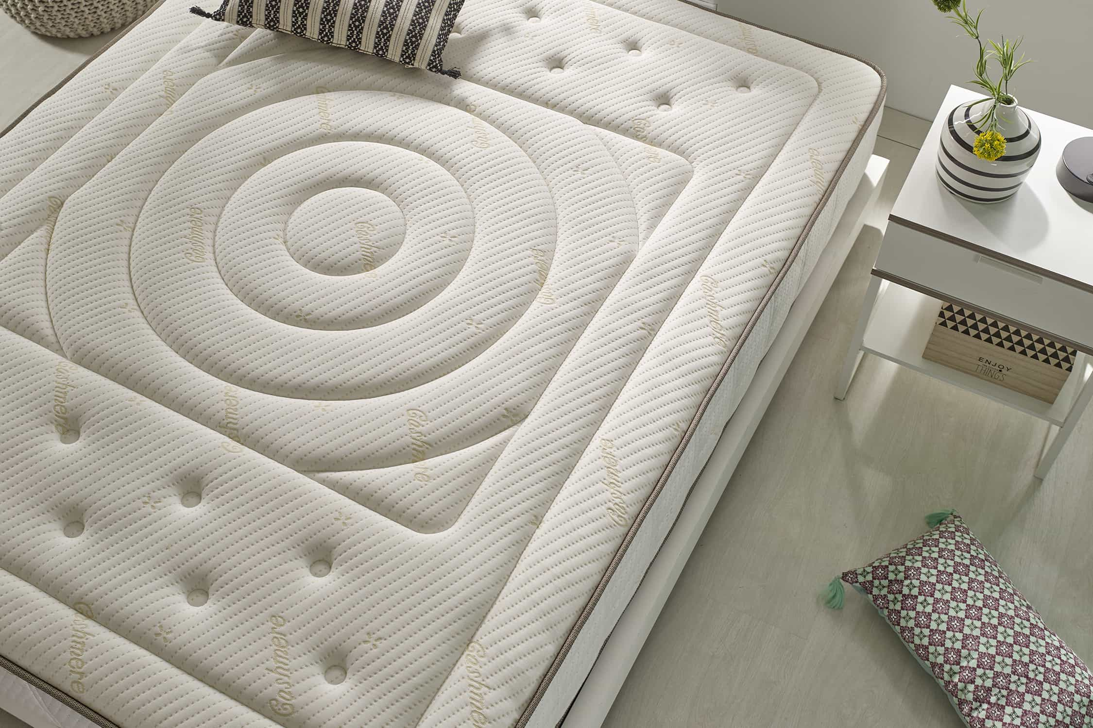 Colchón blanco con diseño en espiral y detalles de botones. Junto a él, una mesa blanca con decoración.